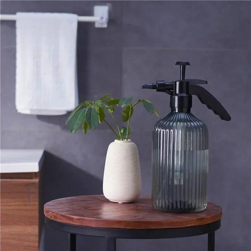 Glass spray bottle and white vase with plant on a wooden table against a gray wall.