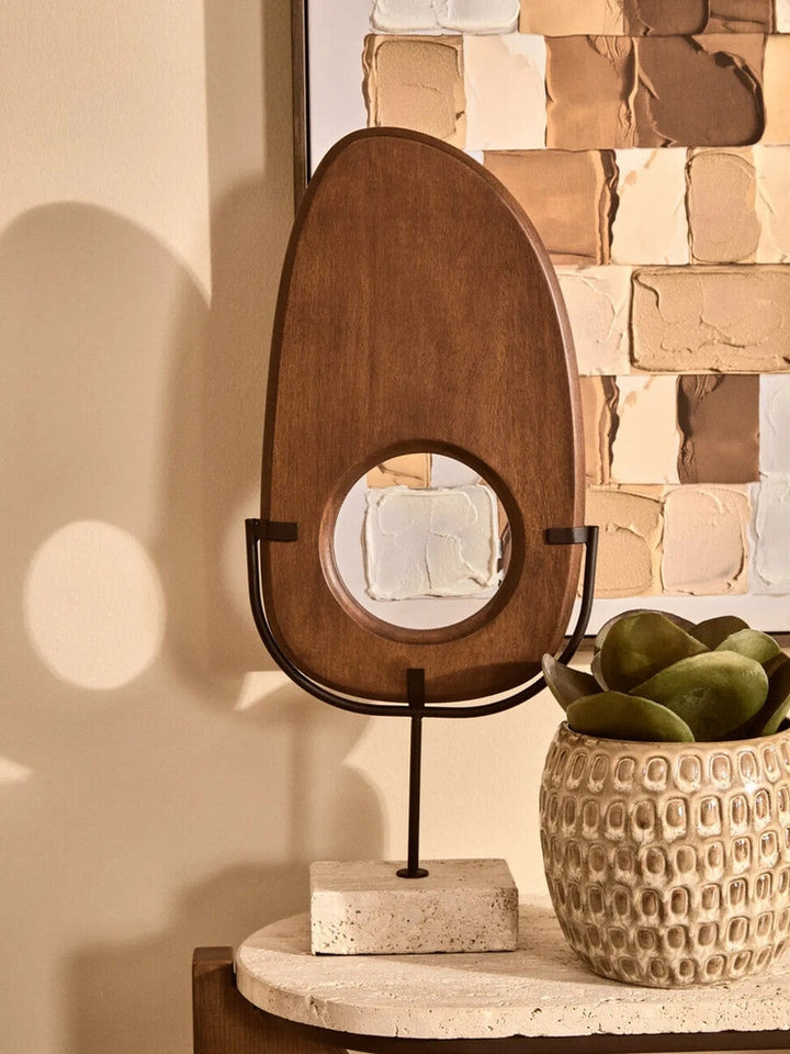 Arsen Wood Vertical Sculpture — oval stained brown wood panel with central circular cutout mounted in a black metal cradle and slender stem on a rectangular light stone base, displayed on a console next to a textured ceramic pot.
