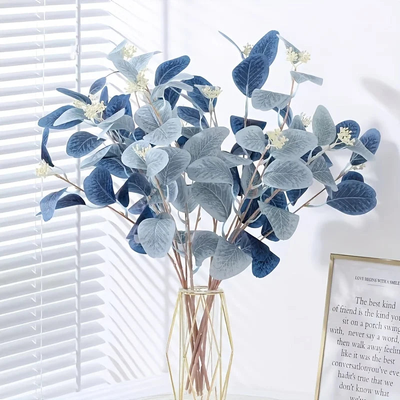 Bouquet of blue and gray artificial leaves in a clear vase against a white wall with a framed quote.