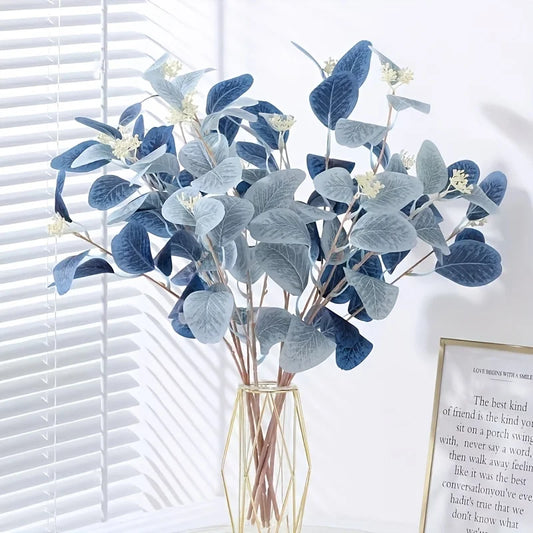Bouquet of blue and gray artificial leaves in a clear vase against a white wall with a framed quote.