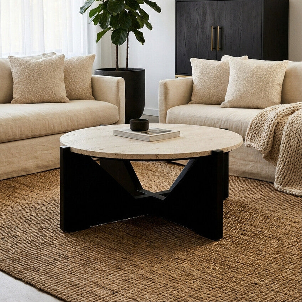 Elsen Travertine Coffee Table with round light travertine top and black geometric wooden base, styled in a neutral living room with beige sofas and jute rug.