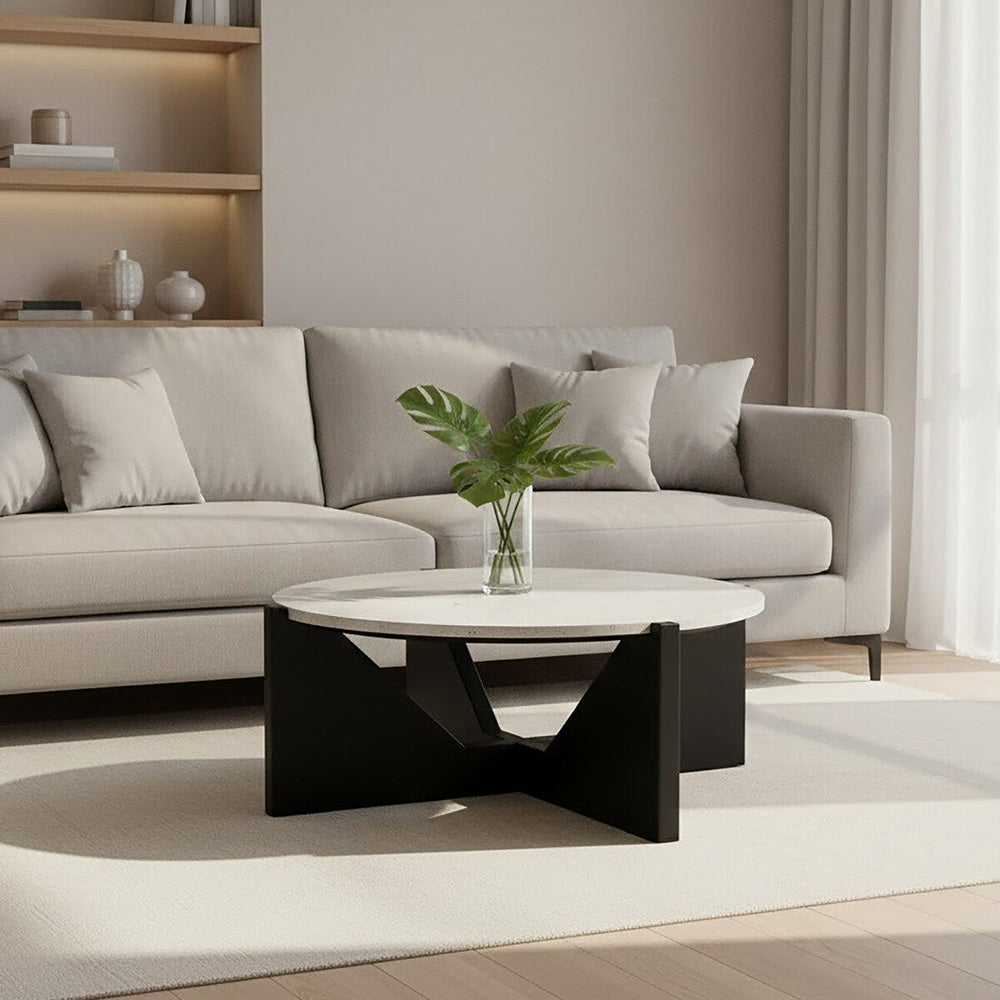 Elsen Travertine Coffee Table — round light travertine top on a black geometric base, styled with a glass vase of green leaves in front of a beige sofa.