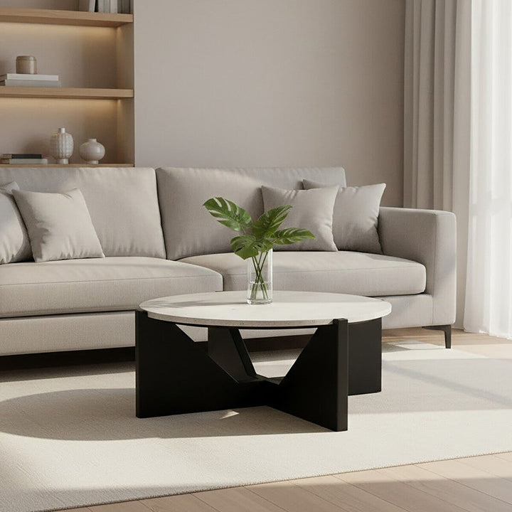 Elsen Travertine Coffee Table — round light travertine top on a black geometric base, styled with a glass vase of green leaves in front of a beige sofa.