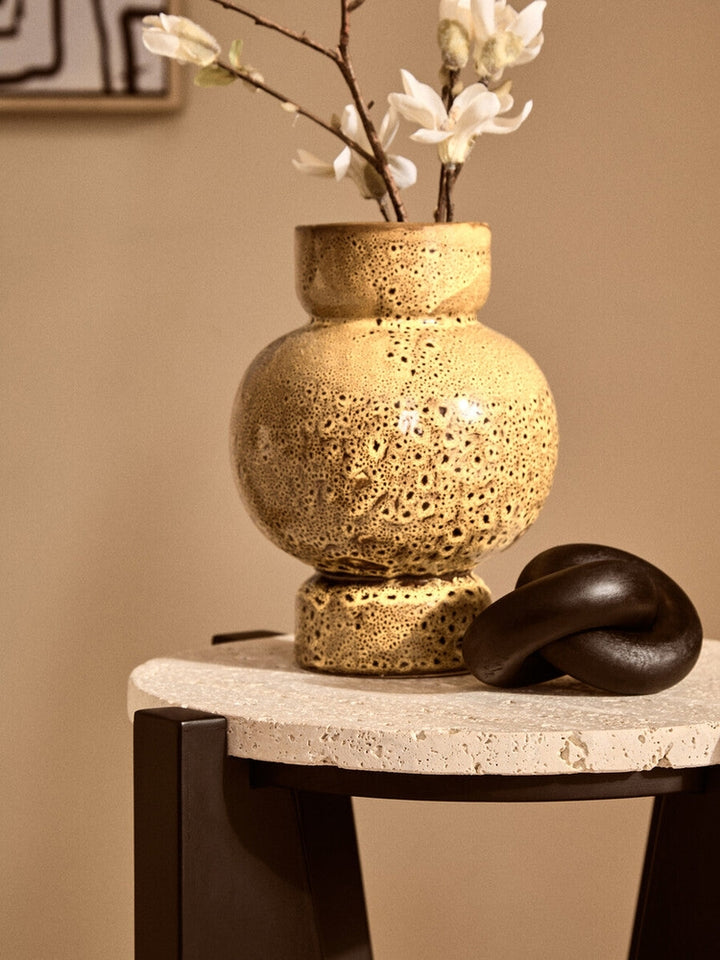 Elsen Travertine Side Table with round porous travertine top and dark legs, styled with a textured yellow ceramic vase holding white blossoms and a black knot decor