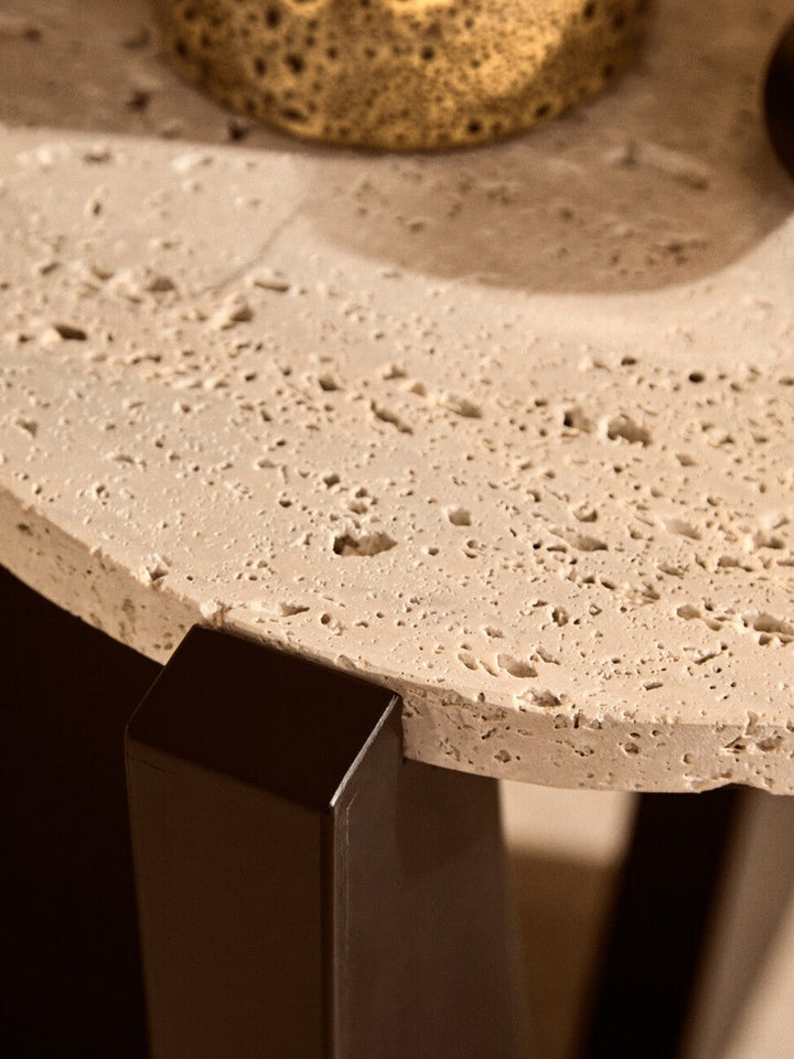 Elsen Travertine Side Table close-up showing round pockmarked travertine tabletop edge and dark rectangular metal leg.