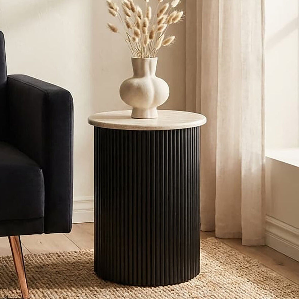 Elsen Travertine Side Table with round light travertine top and black fluted cylindrical base, styled with a sculpted neutral vase and dried grasses.