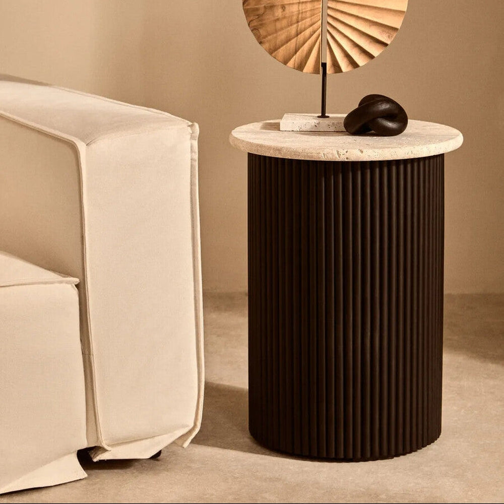 Elsen Travertine Side Table with round light travertine top and ribbed dark cylindrical base, styled beside a cream sofa with a small knot sculpture and fan-shaped tabletop accent.