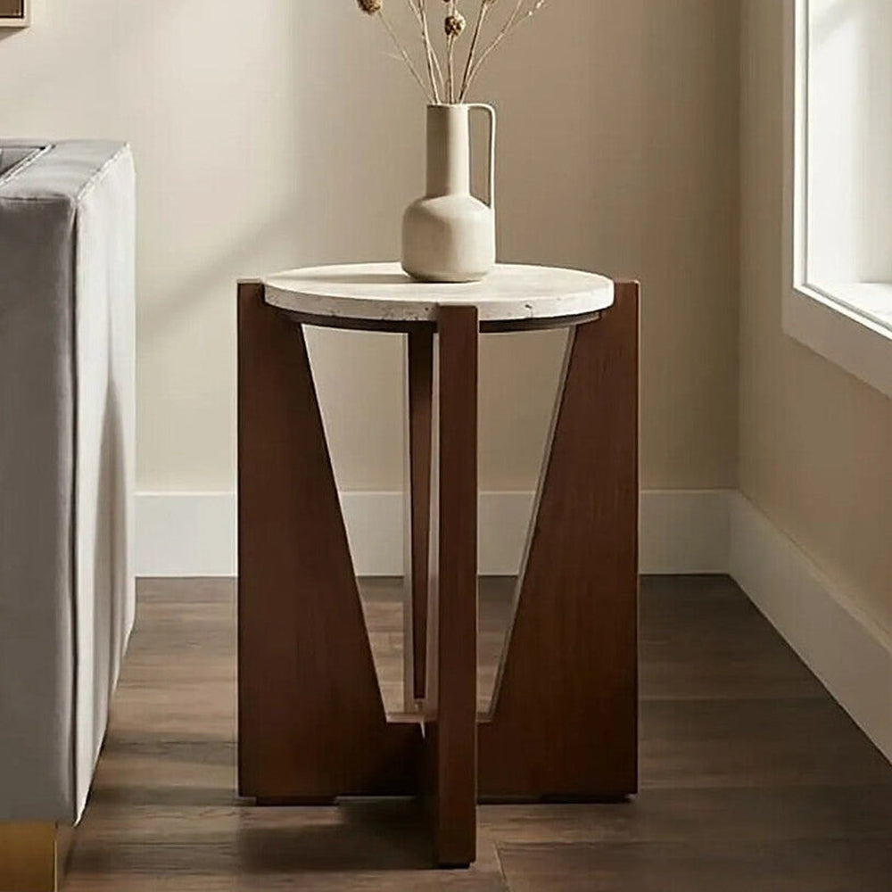Elsen Travertine Side Table with round light travertine top on a warm brown geometric wooden base featuring triangular cutouts, shown beside a sofa.