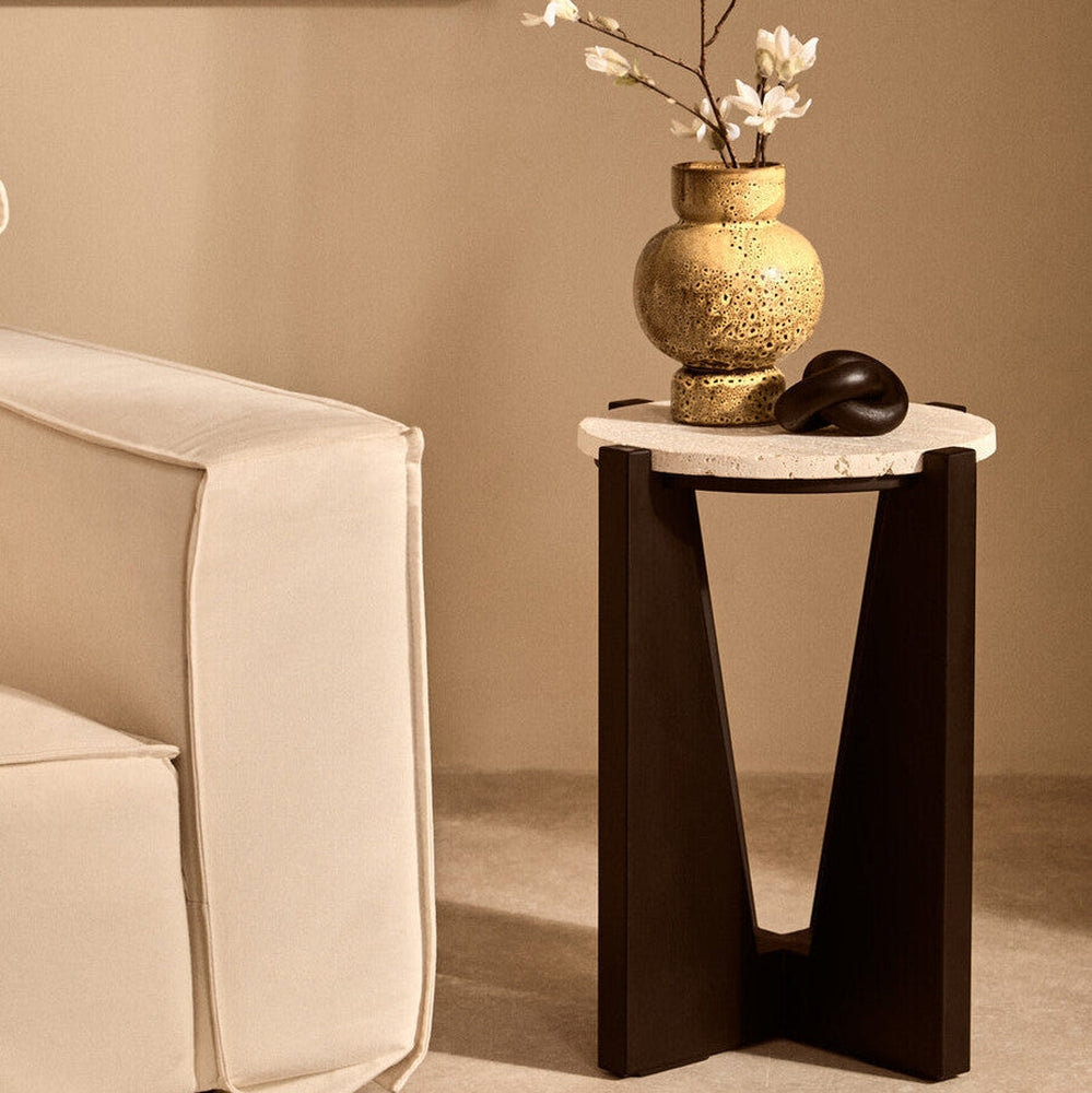 Elsen Travertine Side Table — round light travertine top on a dark V-shaped wooden base, styled beside a cream sofa with a gold textured vase and black knot decor on top.