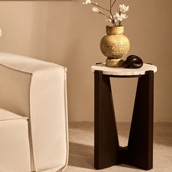 Elsen Travertine Side Table — round light travertine top on a dark V-shaped wooden base, styled beside a cream sofa with a gold textured vase and black knot decor on top.