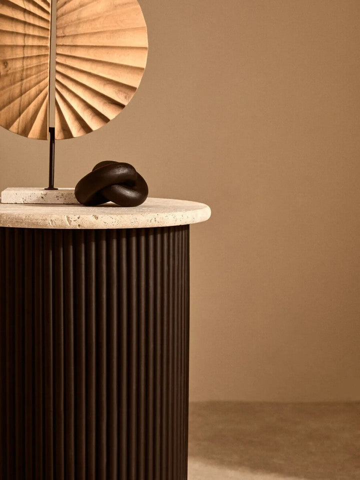 Elsen Travertine Side Table with round light-beige travertine top and dark fluted cylindrical base, styled with a pleated fan and small black knot sculpture