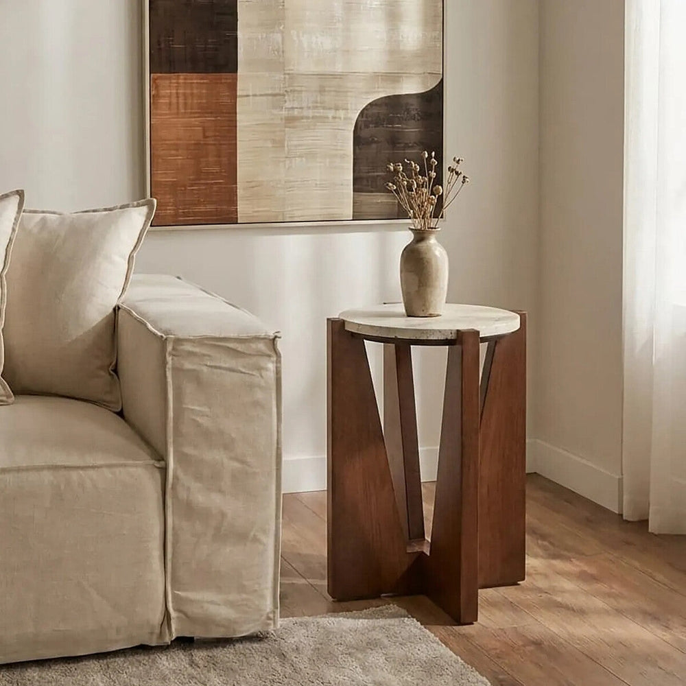 Elsen Travertine Side Table with round light travertine top and geometric brown wooden base, styled beside a beige sofa with a vase of dried stems