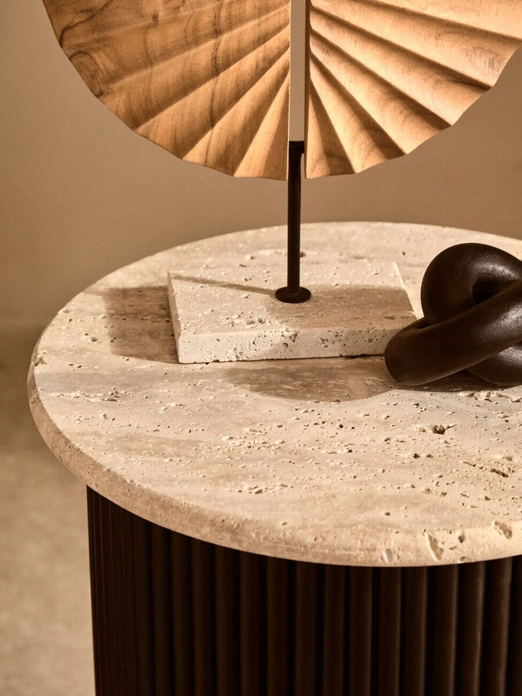 Elsen Travertine Side Table — round textured travertine top with natural pitting and a dark fluted cylindrical base, shown styled with small sculptural decor on top