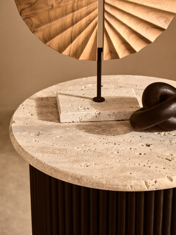 Elsen Travertine Side Table — round textured travertine top with natural pitting and a dark fluted cylindrical base, shown styled with small sculptural decor on top