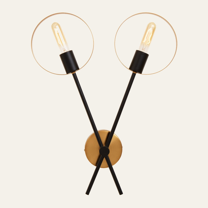 Erathia Two Bulb Ring Wall Light with two crossed black rods, exposed filament bulbs in black sockets framed by thin gold rings, mounted on a round gold wall plate