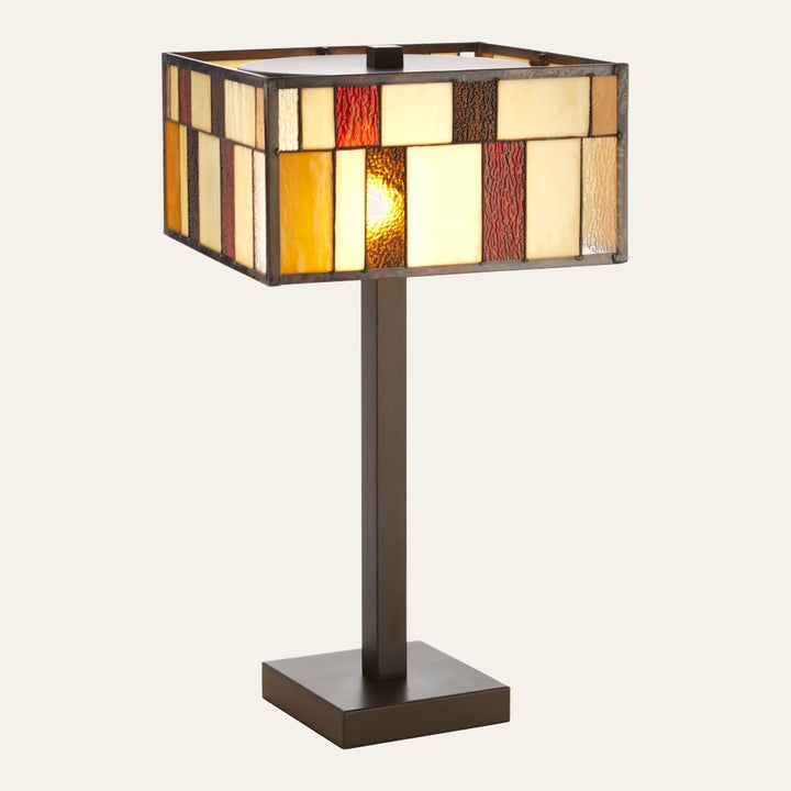 Evaya Table Lamp with rectangular stained-glass mosaic shade in cream, amber and red panels, dark metal square stem and base, warm glowing bulb visible through textured glass