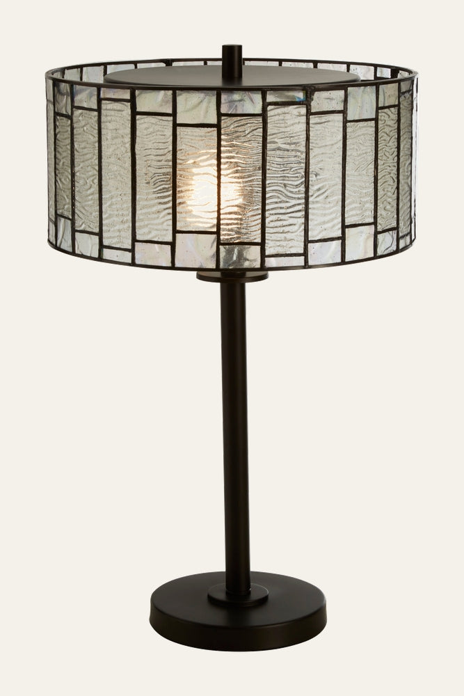 Evaya Table Lamp with drum stained-glass shade of textured rectangular clear glass panels, lit bulb inside, and slim dark metal rod with round base