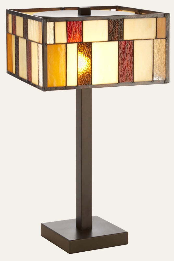 Evaya Table Lamp with rectangular stained-glass shade of amber, cream and red textured glass panels, lit on a dark bronze square metal stem and base.