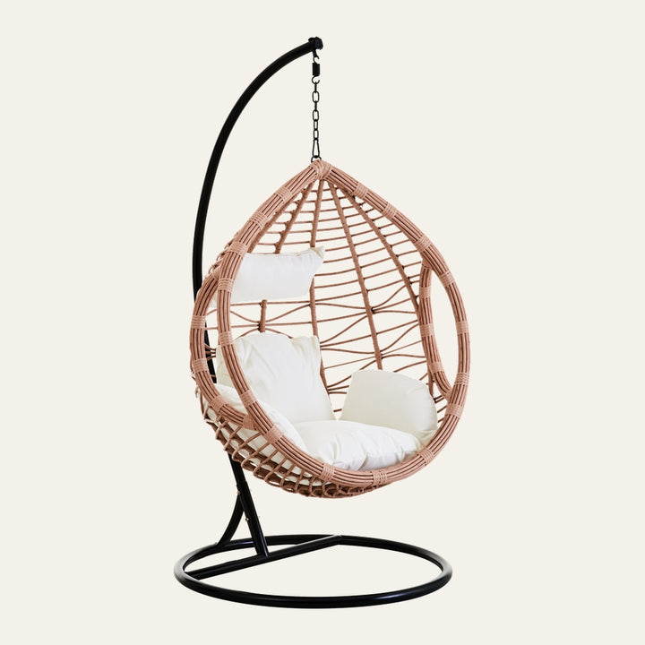 Evira Rattan Effect Hanging Chair with egg-shaped light tan rattan-effect weave suspended from a black metal stand with circular base, fitted with white seat cushion and pillows.