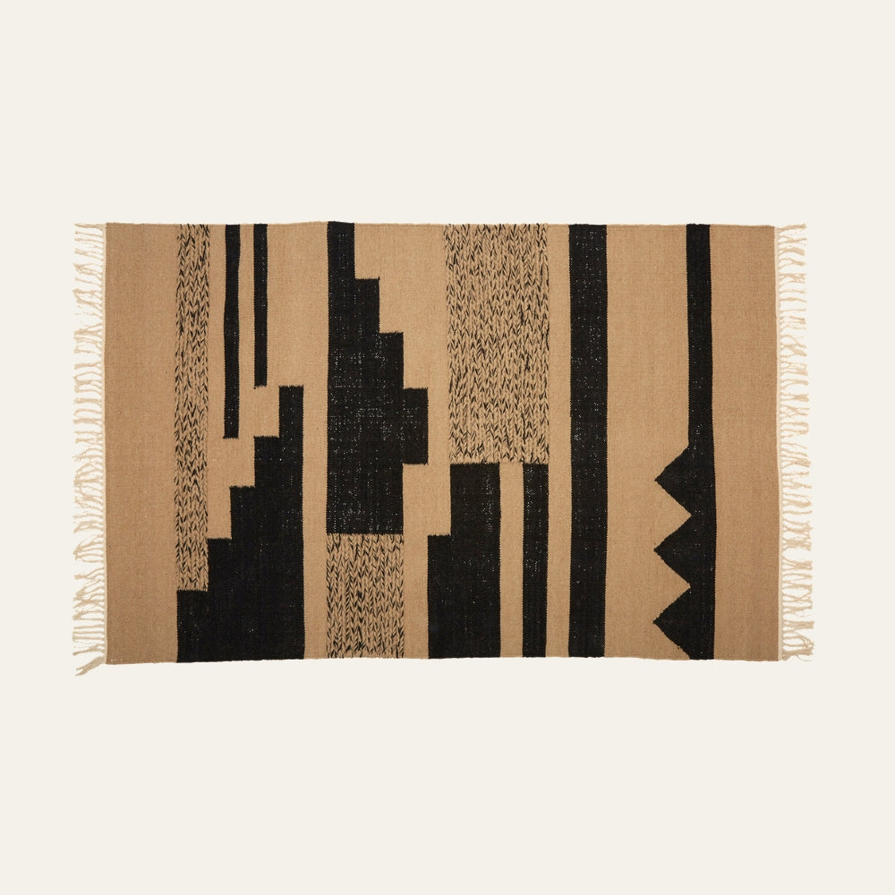 Ezelle Trenza Fabric Rug — rectangular woven fabric rug in tan with bold black geometric blocks, vertical stripes and textured motifs, short fringed edges