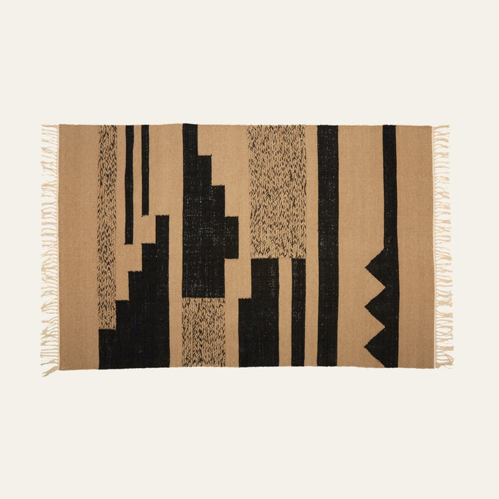 Ezelle Trenza Fabric Rug — rectangular woven fabric rug in tan with bold black geometric blocks, vertical stripes and textured motifs, short fringed edges