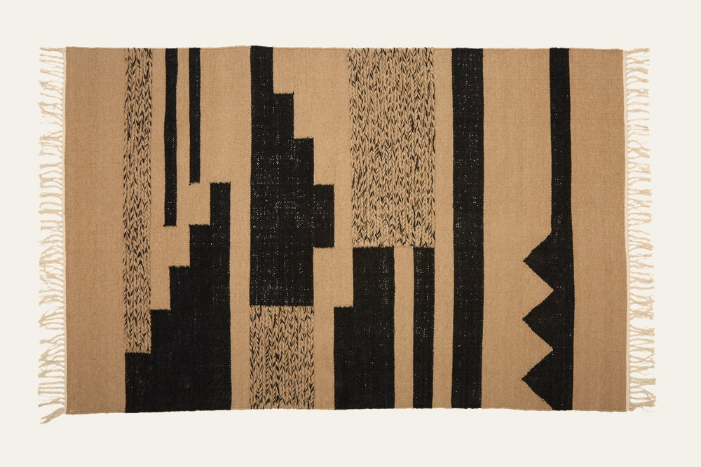 Ezelle Trenza Fabric Rug — rectangular tan woven fabric with bold black geometric vertical stripes and blocks, textured weave and fringe tassel edges.