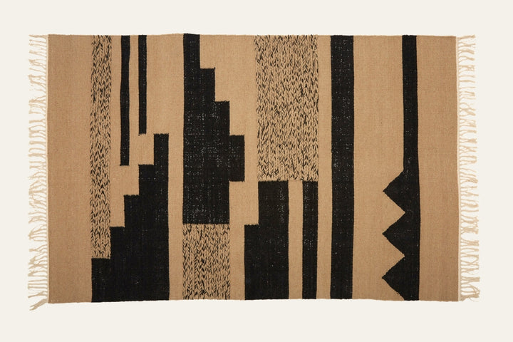 Ezelle Trenza Fabric Rug — rectangular tan woven fabric with bold black geometric vertical stripes and blocks, textured weave and fringe tassel edges.