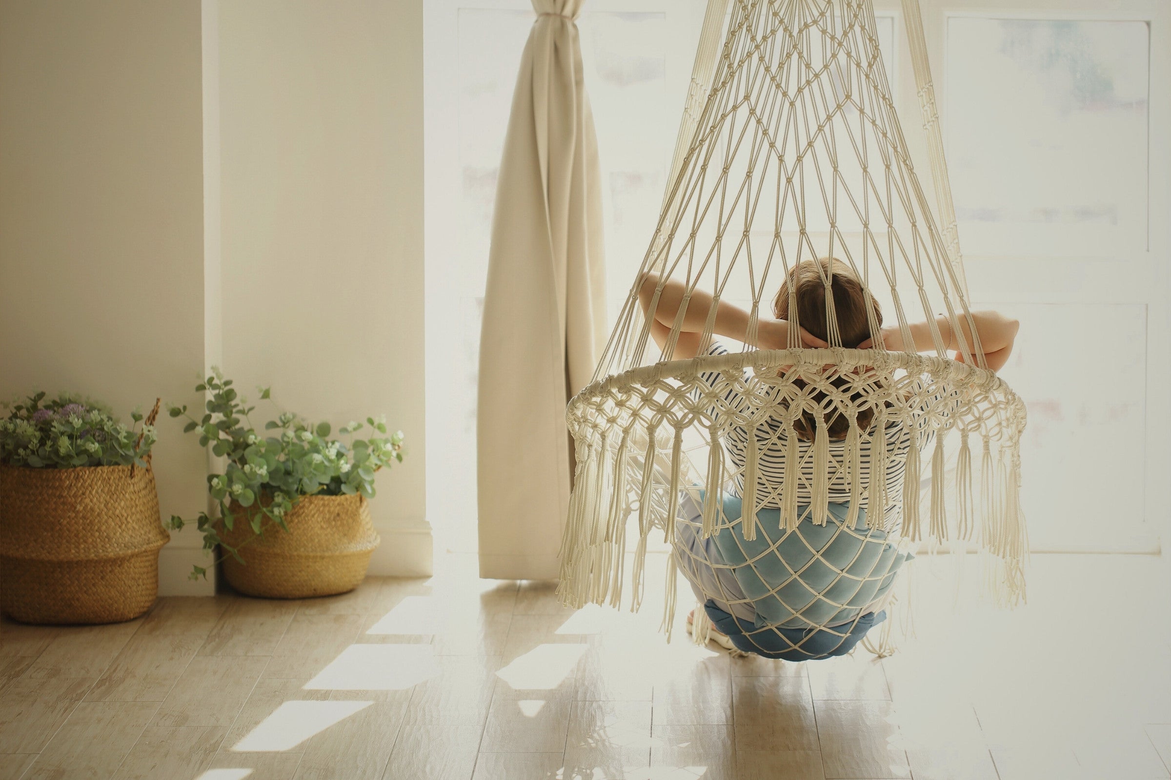 Person sitting in a macrame hammock in a bright room with plants and curtains.