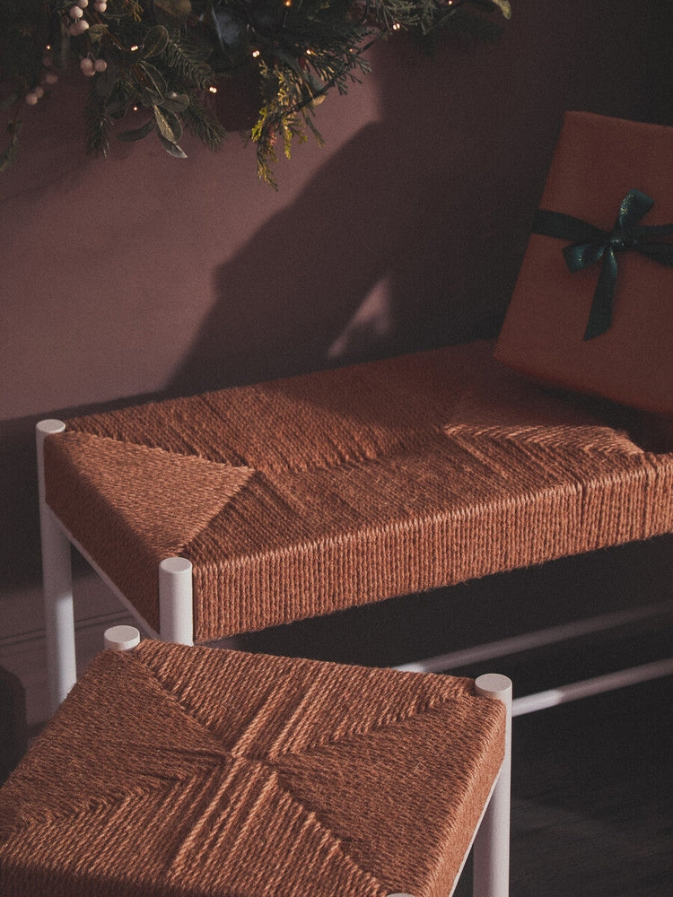Quildor Open Frame Bench — white open metal frame with woven brown fiber seat, shown with a matching woven stool and holiday greenery