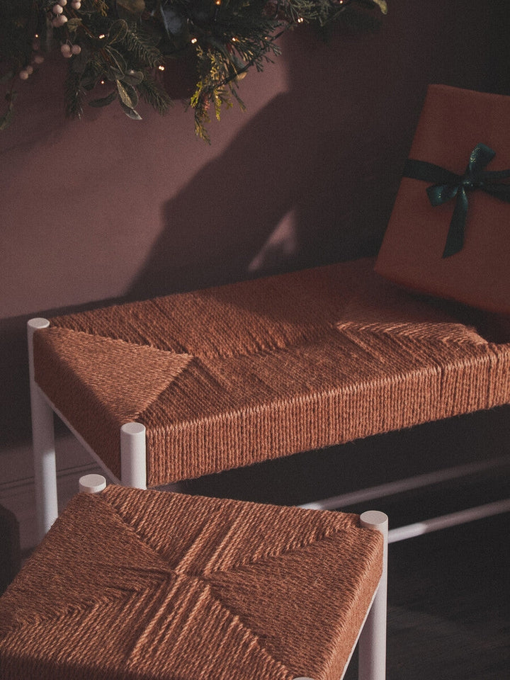 Quildor Open Frame Bench — white open metal frame with woven brown fiber seat, shown with a matching woven stool and holiday greenery