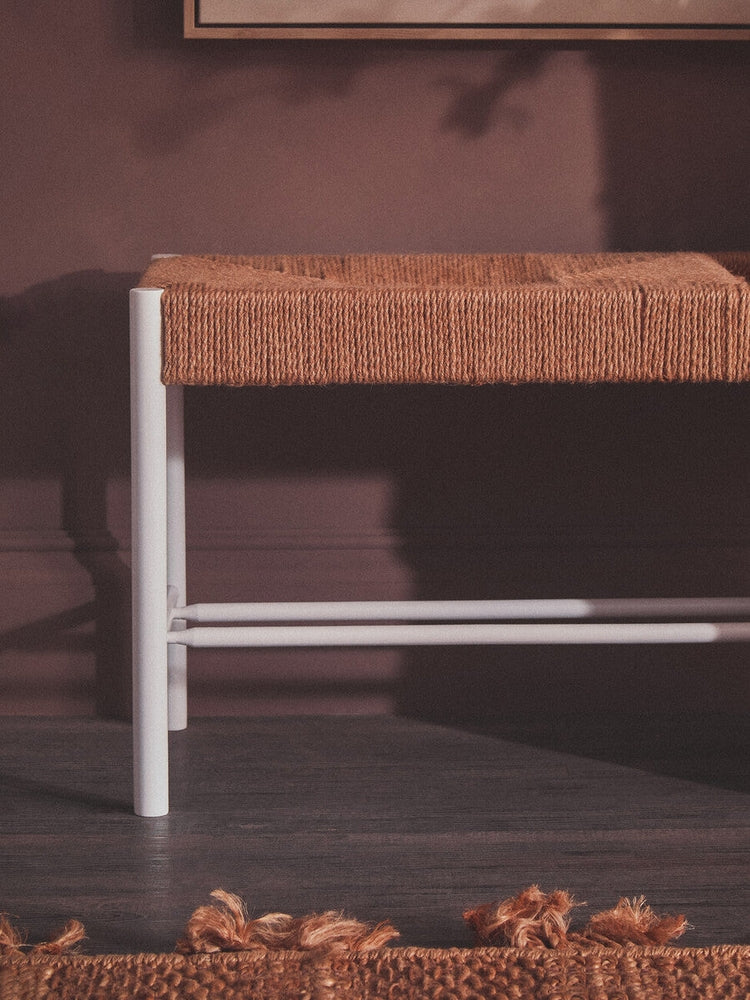 Quildor Open Frame Bench with woven tan rope seat and white open metal frame, pictured against a mauve wall on a dark floor with a fringed rust-colored rug