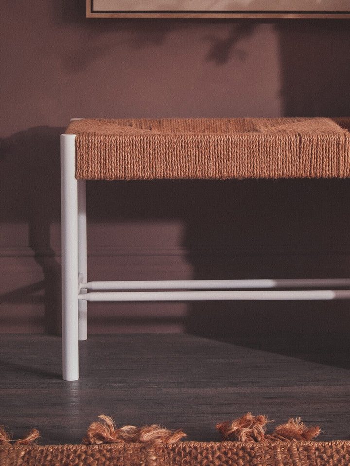 Quildor Open Frame Bench with woven tan rope seat and white open metal frame, pictured against a mauve wall on a dark floor with a fringed rust-colored rug