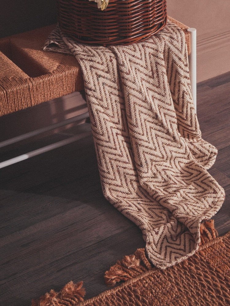 Quildor Open Frame Bench with a woven natural-fiber seat and white metal open frame, draped with a beige chevron knit throw and a wicker basket on top.