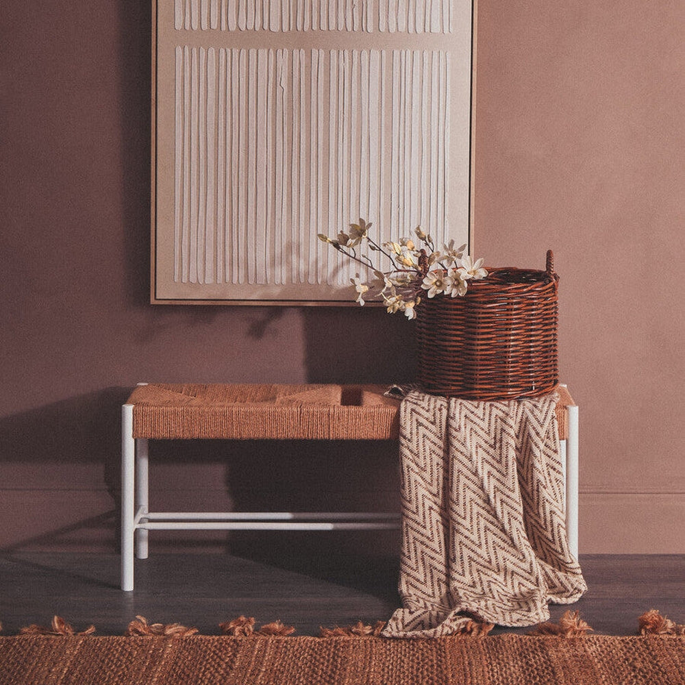 Quildor Open Frame Bench with tan woven seat and white metal open frame, styled with a wicker basket of flowers and a chevron-patterned throw against a muted mauve wall.