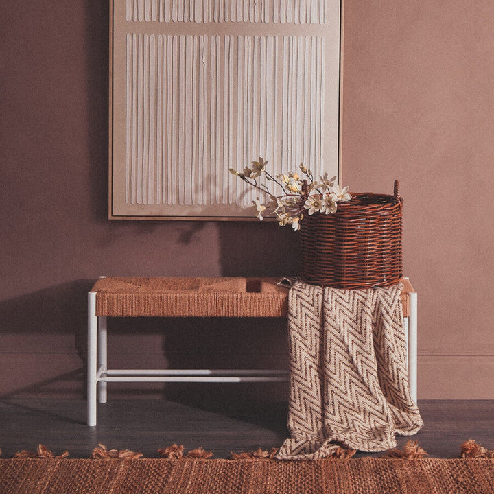 Quildor Open Frame Bench with tan woven seat and white metal open frame, styled with a wicker basket of flowers and a chevron-patterned throw against a muted mauve wall.