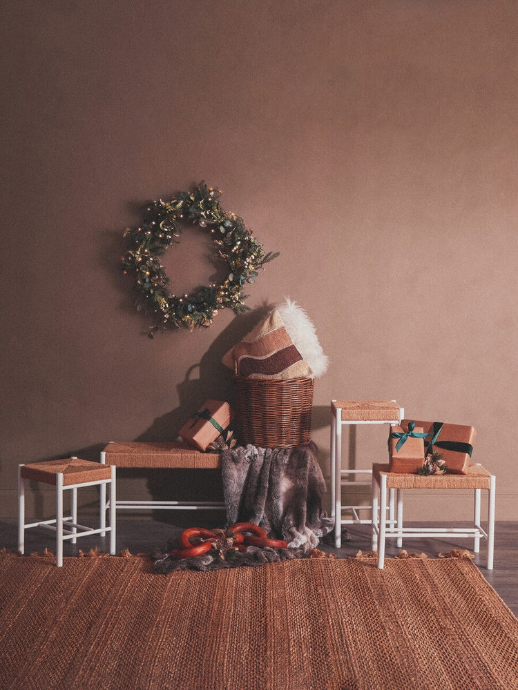 Quildor Open Frame Bench with white metal open frame and woven natural-fiber seat, styled in a holiday vignette with a wicker basket, fur throw, pillows, wreath and wrapped gifts