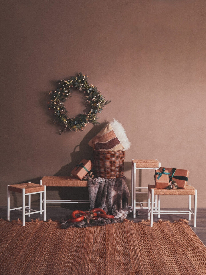 Quildor Open Frame Bench with white metal open frame and woven natural-fiber seat, styled in a holiday vignette with a wicker basket, fur throw, pillows, wreath and wrapped gifts