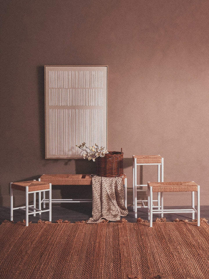 Quildor Open Frame Bench with white metal open frame and woven natural-fiber seat, styled in a neutral room with matching stools, wicker basket, patterned throw and textured wall art.