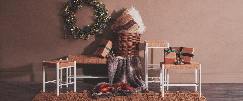 Quildor Open Frame Bench with white open metal frame and woven natural-fiber seat, styled with a wreath, wrapped gifts, wicker basket and faux fur throw on a jute rug.