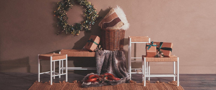Quildor Open Frame Bench with white open metal frame and woven natural-fiber seat, styled with a wreath, wrapped gifts, wicker basket and faux fur throw on a jute rug.