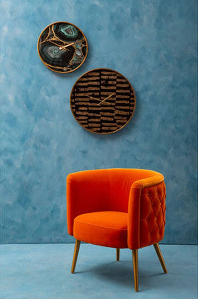 Soren Wall Clock: two round wall clocks with gold-tone hands and rims — one teal agate-style face and one brown textured striped face — mounted on a blue textured wall above an orange velvet chair