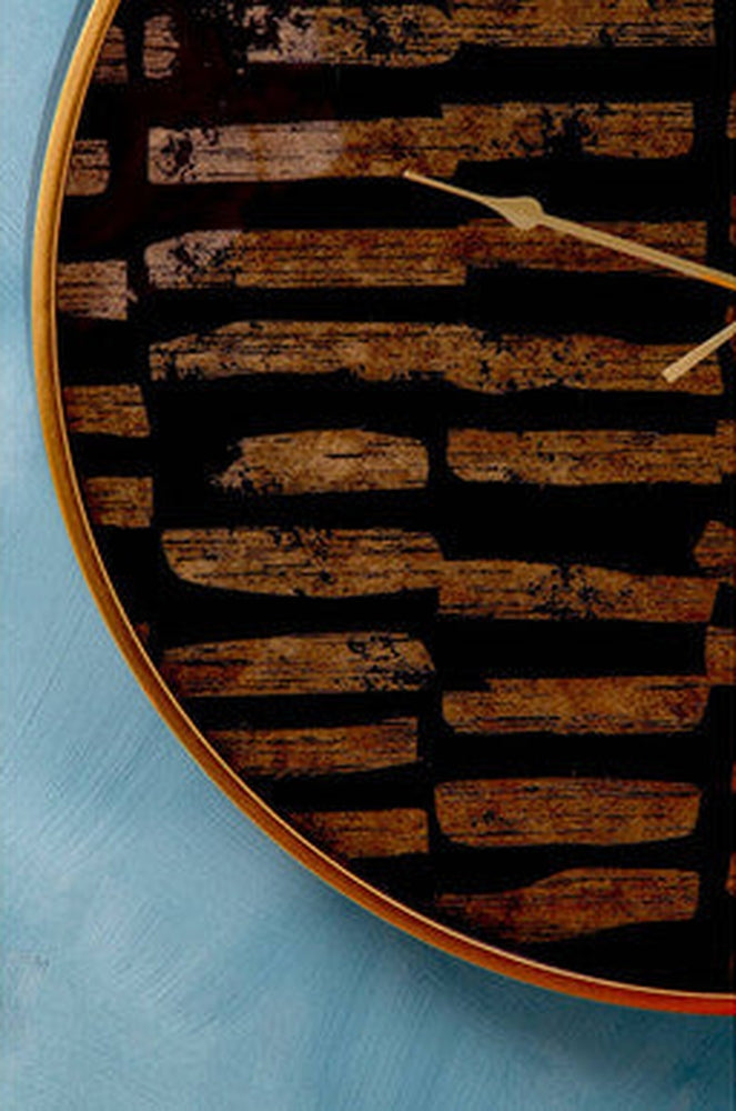 Soren Wall Clock close-up: round clock with black face patterned in distressed brown wood-plank stripes, thin gold bezel and slim gold hands against blue and cream wall