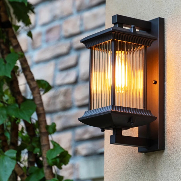 Black outdoor wall light glowing warmly beside a stone wall and green foliage